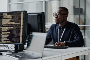 A software developer or cybersecurity analyst wearing glasses and a lanyard sits at a multi-monitor workstation displaying lines of code, working in a modern office environment